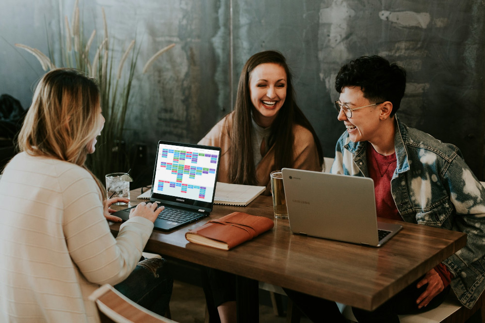 A team of three collaborating joyfully at a shared desk with a digital kanbantool.com board visible