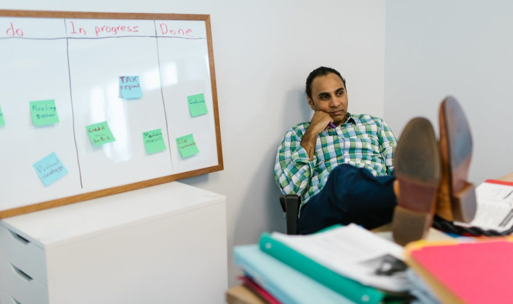 Man deep in thought sitting at a desk with a simple Kanban board behind him